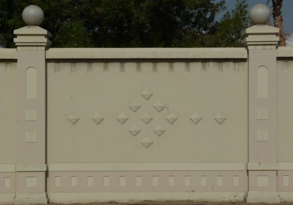 Concrete fence texture made up of a solid tan wall with a diamond pattern running through the middle, and a ball-topped stone column on either side. Rectangular recessions run along the top and bottom of the fence, and up the columns.